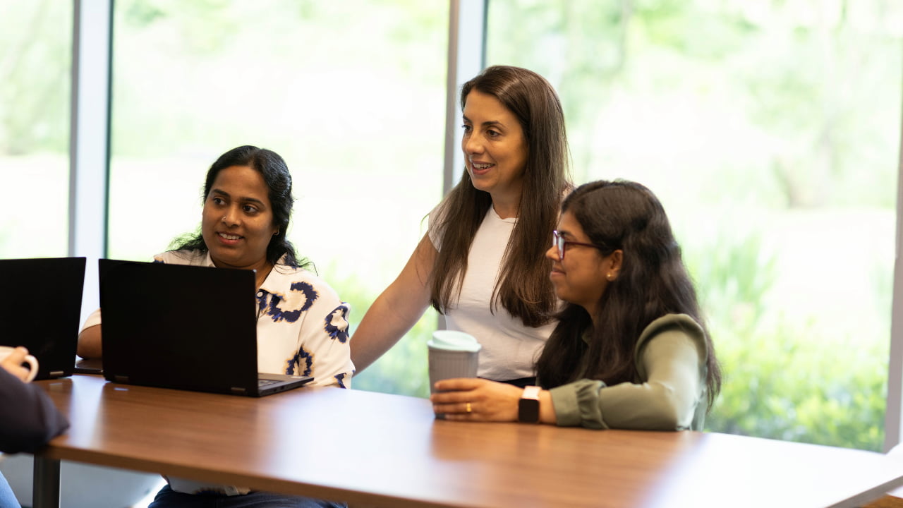 Three employees sitting around a laptop, one is holding coffee