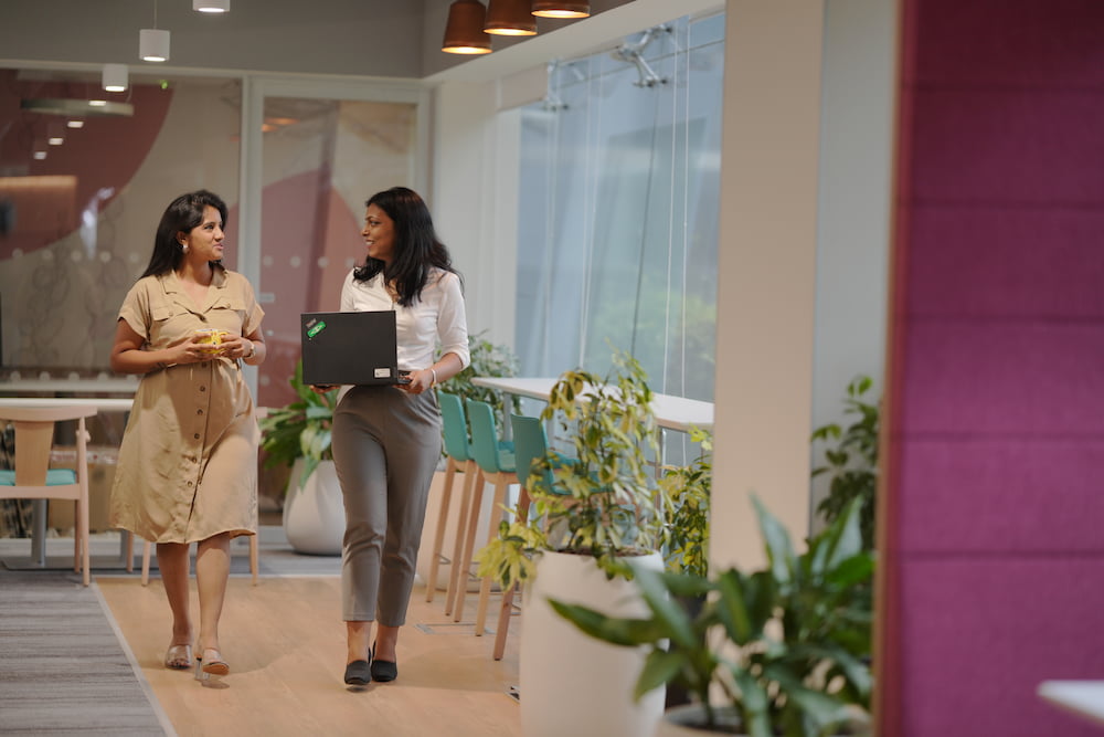 Two women walking together and talking in an office. One is holding a laptop