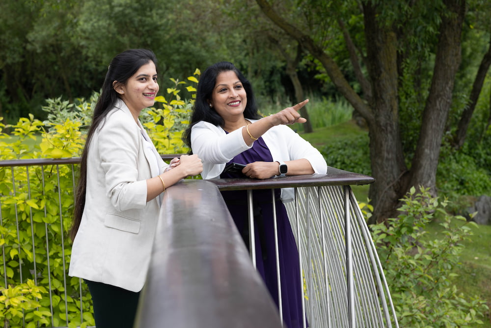 Two women standing at a railing near a lush outdoor environment. One is pointing at something