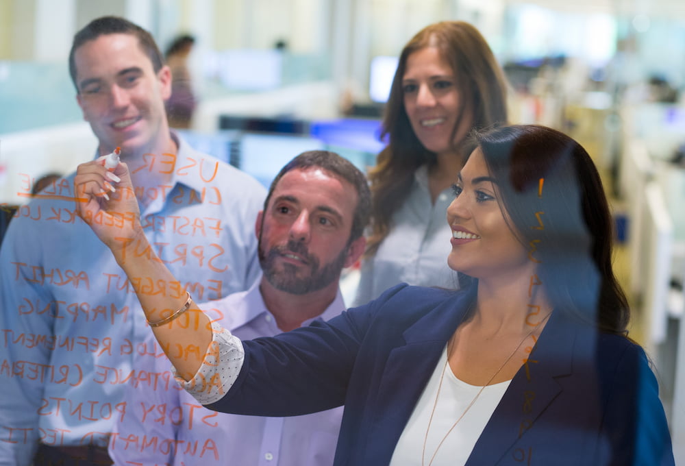 A woman is writing on a clear surface with an orange marker while a group of coworkers watch her